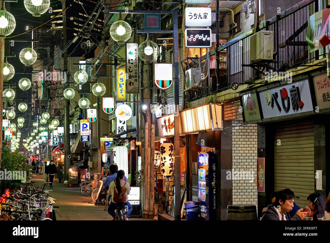 Des lampes globes pendent au-dessus d'une ruelle de restaurant étroite avec des lanternes kushikatsu et des vélos garés, tandis que les gens dînent et font du vélo par une soirée de printemps. Banque D'Images