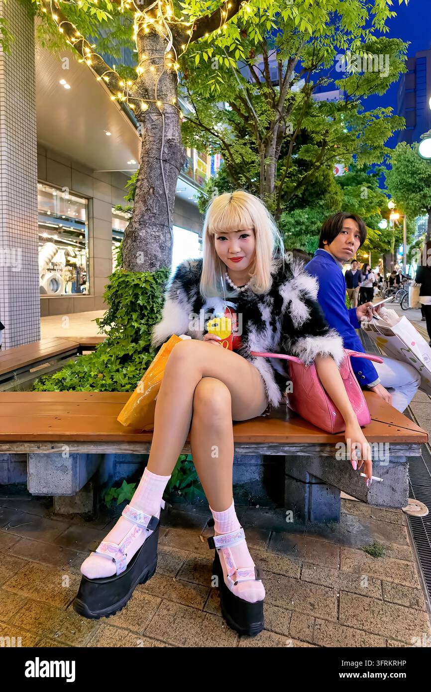Femme blonde en veste en fausse fourrure, chaussettes roses et sandales à plateformes fumant sur un banc au crépuscule au Japon, homme avec téléphone à côté d'elle. Banque D'Images