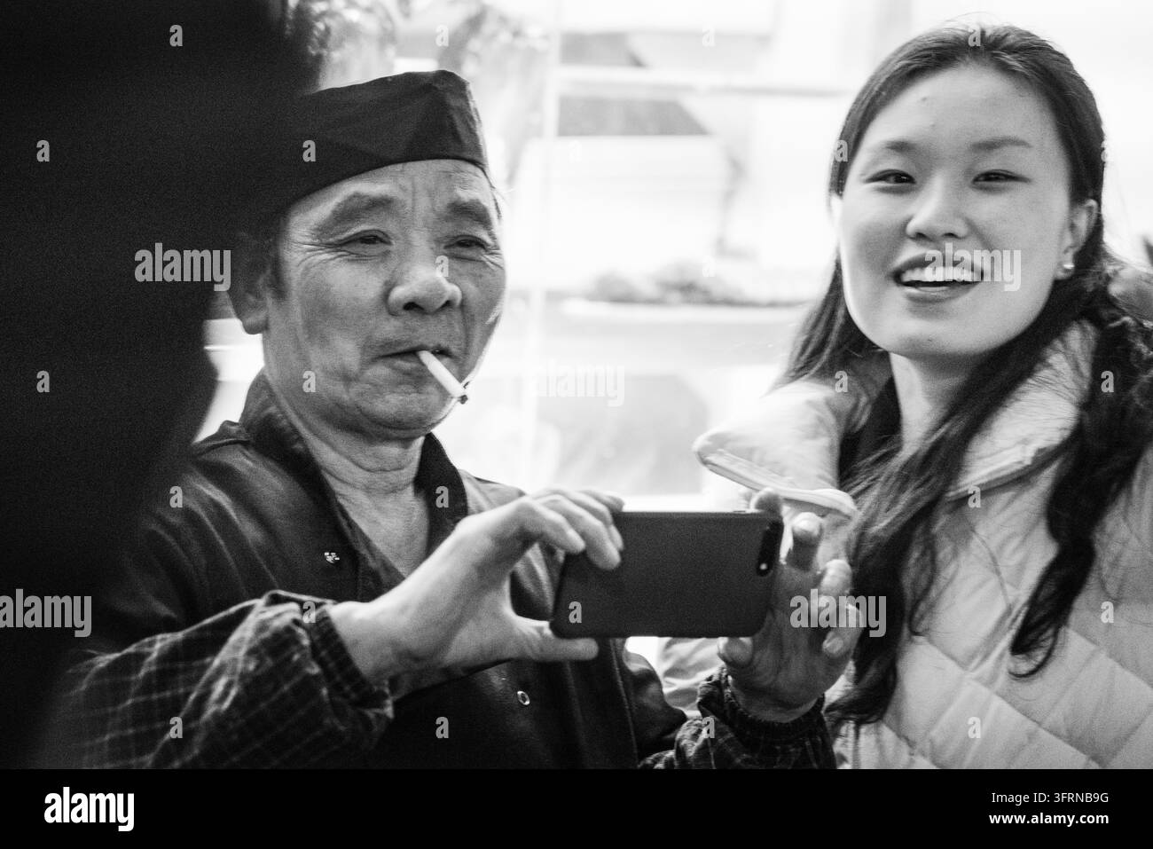 HOMME ET FEMME REGARDENT L'ÉCRAN DU TÉLÉPHONE, CHINATOWN DE LONDRES EN HIVER, REPORTAGE DE VOYAGE, LONDRES, ANGLETERRE, 2019 : Sur une photographie en noir et blanc, un homme avec une cigarette dans la bouche tient un téléphone tandis qu'une jeune femme souriante regarde vers l'appareil photo. INFO : Le Chinatown de Londres, niché au cœur vibrant de Soho, est un centre culturel animé, célèbre pour ses restaurants asiatiques authentiques, ses boulangeries traditionnelles et ses rues éclairées au néon. Les lanternes rouges emblématiques offrent un arrière-plan parfait pour les amateurs de gastronomie. Banque D'Images