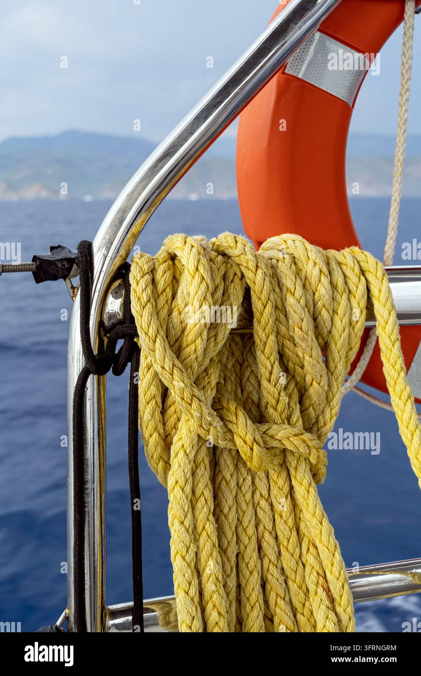 gros plan d'une bobine de corde nautique jaune fixée à une rambarde en acier inoxydable à côté d'un bouée de sauvetage orange sur un yacht, avec une mer bleue calme et une côte lointaine Banque D'Images