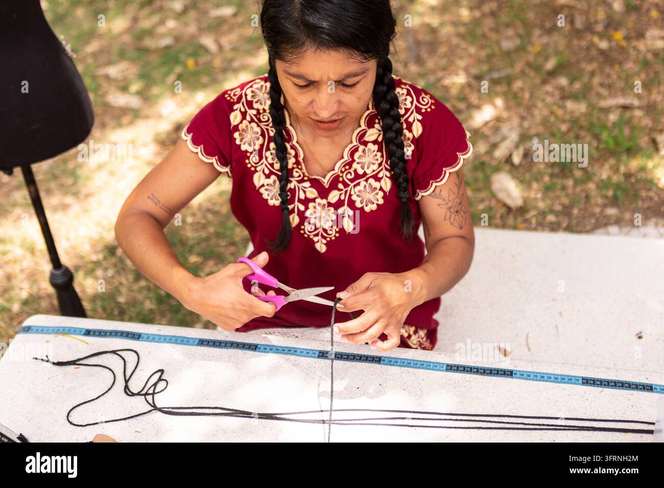 Vue aérienne d'un artisan vêtu de vêtements brodés coupant du cordon pour créer des objets en macramé artisanaux à une table d'extérieur, mettant en valeur des textiles traditionnels Banque D'Images