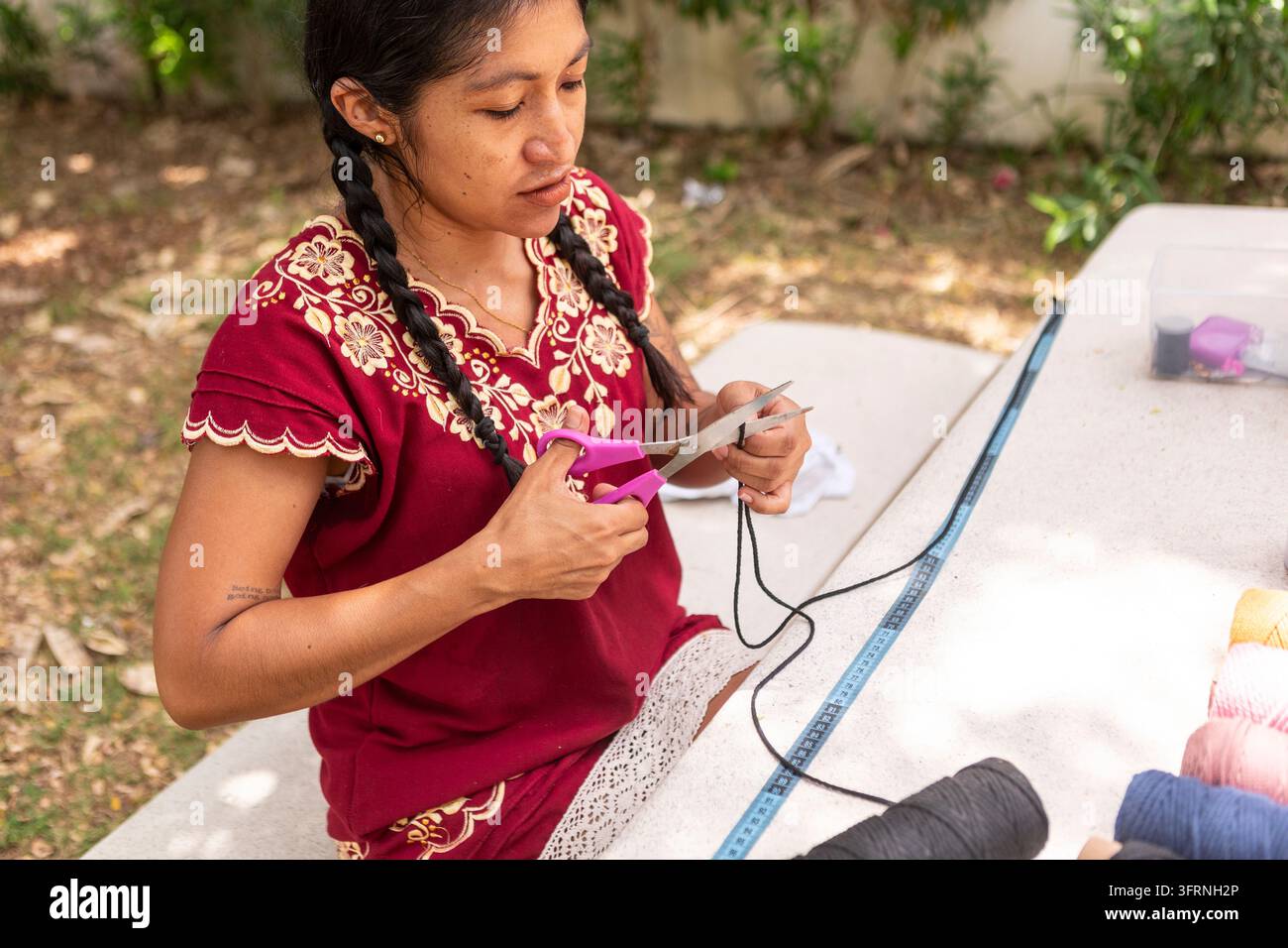 Une femme artisan utilise des ciseaux et une corde pour créer des textiles en macramé artisanaux à une table d'extérieur, pratiquant l'artisanat traditionnel et le travail créatif de loisir Banque D'Images