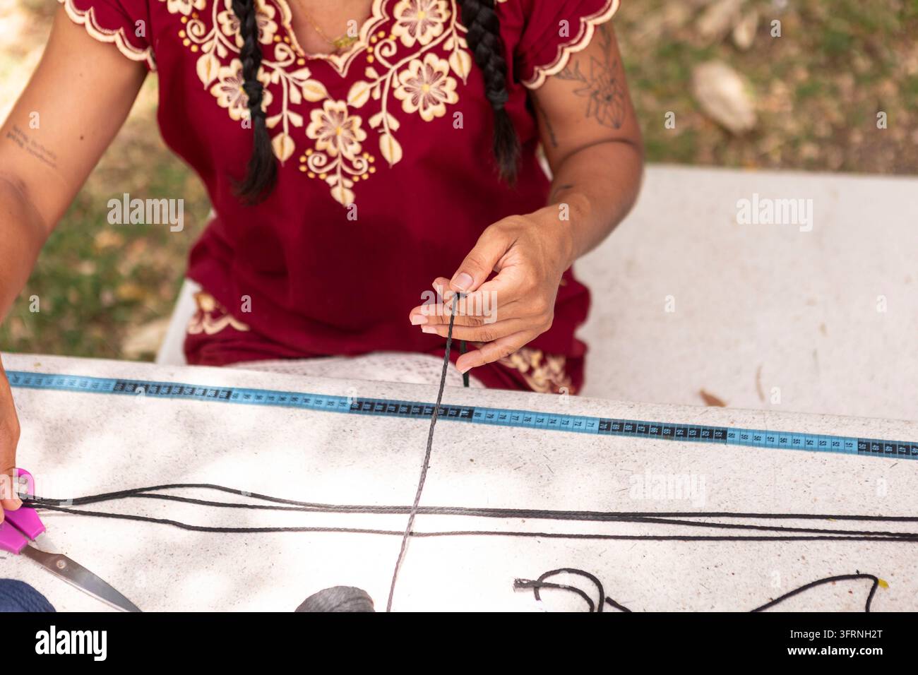 Vue rapprochée d'un artisan vêtu de vêtements brodés mesurant et coupant du cordon noir pour créer des textiles en macramé artisanaux à une table extérieure. Banque D'Images