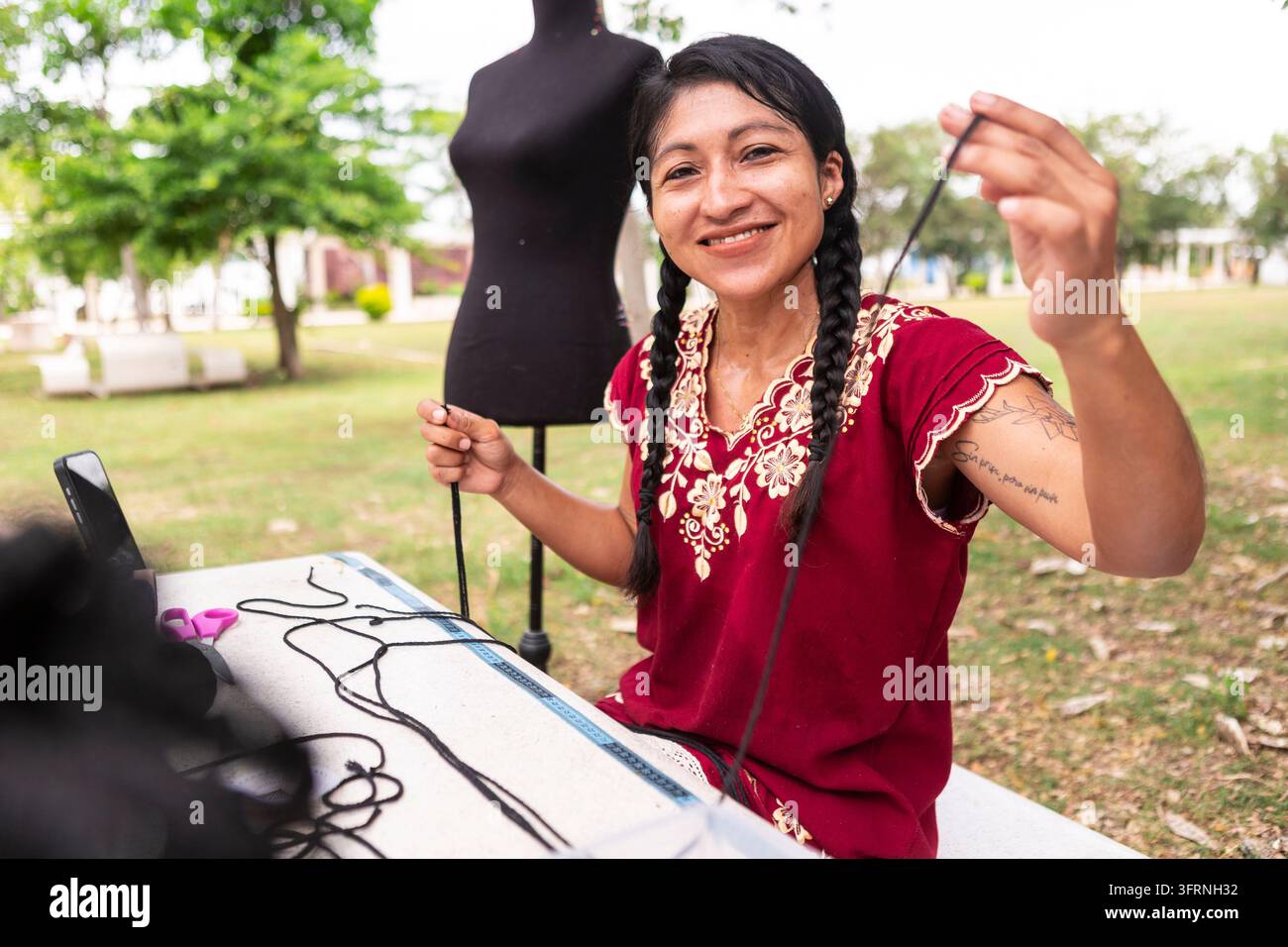 Femme artisan souriante présente un cordon noir tout en créant un textile en macramé fait main à une table extérieure, avec un mannequin et des outils de bricolage dans un parc Banque D'Images