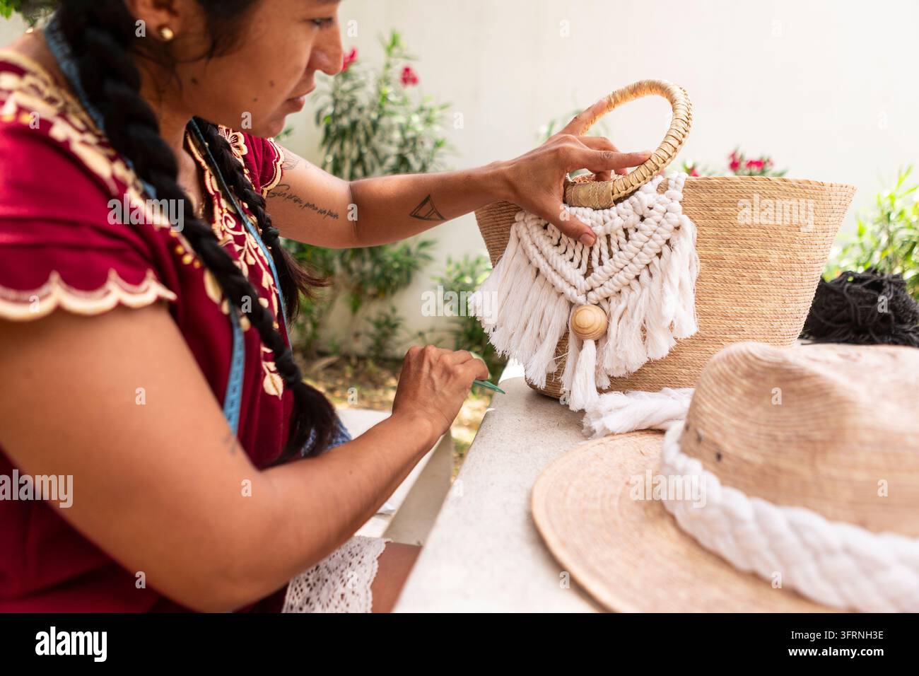 Un artisan crée un sac en macramé fait main avec une frange tissée et une poignée en bois à une table d'artisanat en plein air, entouré de chapeaux de paille, de fil et d'accessoires textiles Banque D'Images