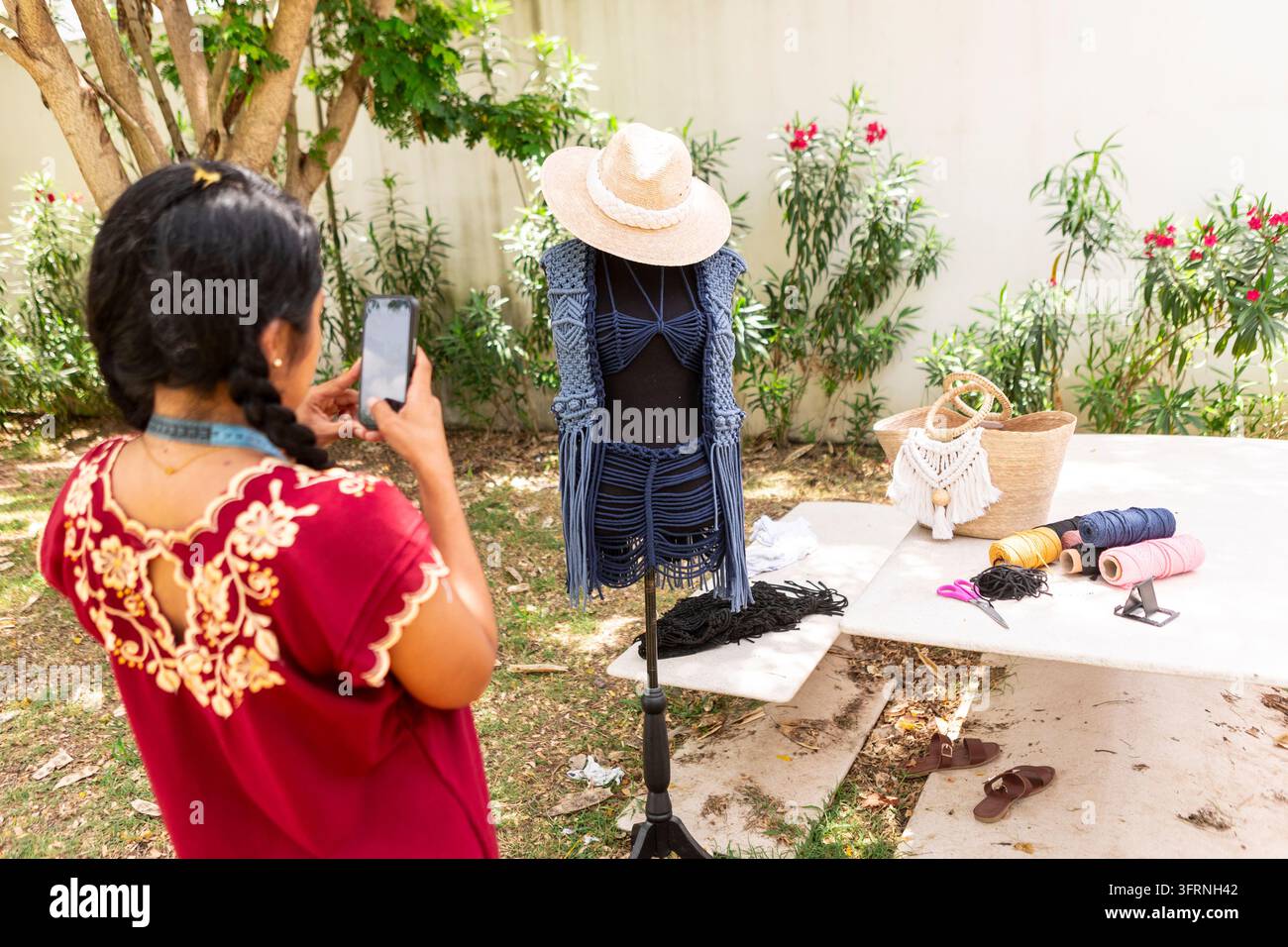 Artisan photographie un vêtement en macramé fait à la main exposé sur un mannequin dans un jardin ensoleillé, avec des cordes textiles, des outils d'artisanat et des accessoires tissés arra Banque D'Images