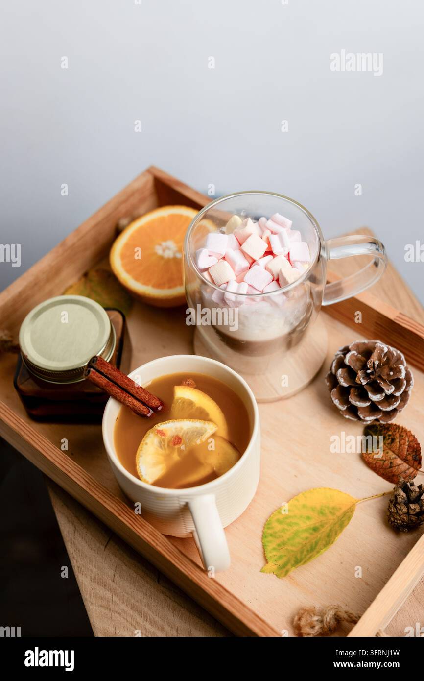 Arrangement de boissons automnales confortables avec thé aux agrumes, cannelle, pot de miel et guimauve, servi sur un plateau en bois à côté d'une pomme de pin et de feuilles colorées. Banque D'Images