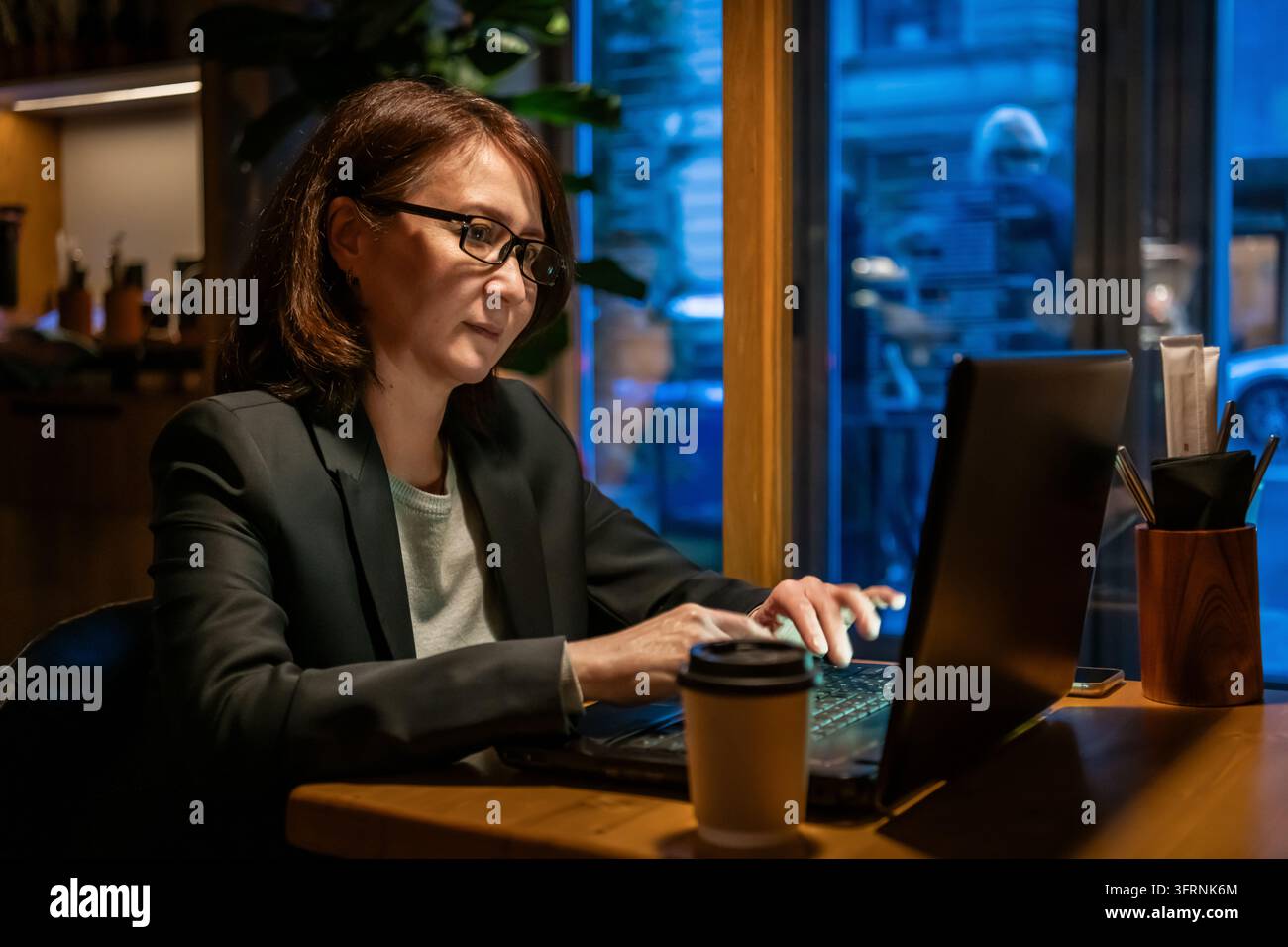 Femme mûre portant des lunettes travaillant sur un ordinateur portable à une table en bois dans un café confortable, éclairée par une lumière intérieure chaude et des reflets bleus de fenêtre. Banque D'Images