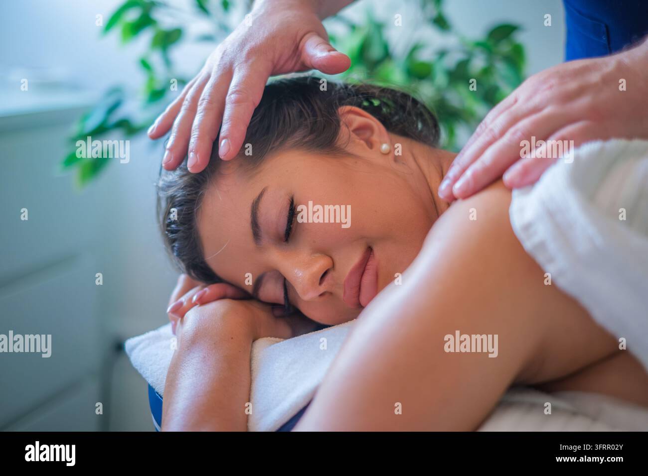 Une jeune femme sereine reposant sur une table de massage les yeux fermés, vivant un massage des épaules profondément relaxant par un thérapeute professionnel Banque D'Images