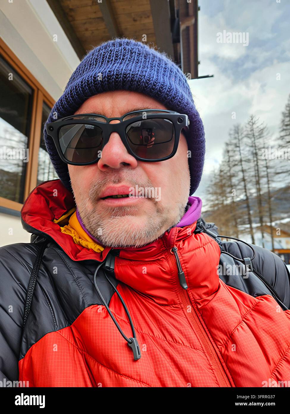 Un homme portant un bonnet tricoté bleu, des lunettes de soleil et une deuxième paire de lunettes avec caméra se tient devant un bâtiment. Il porte une veste rouge et une écharpe violette dans une station de ski Banque D'Images
