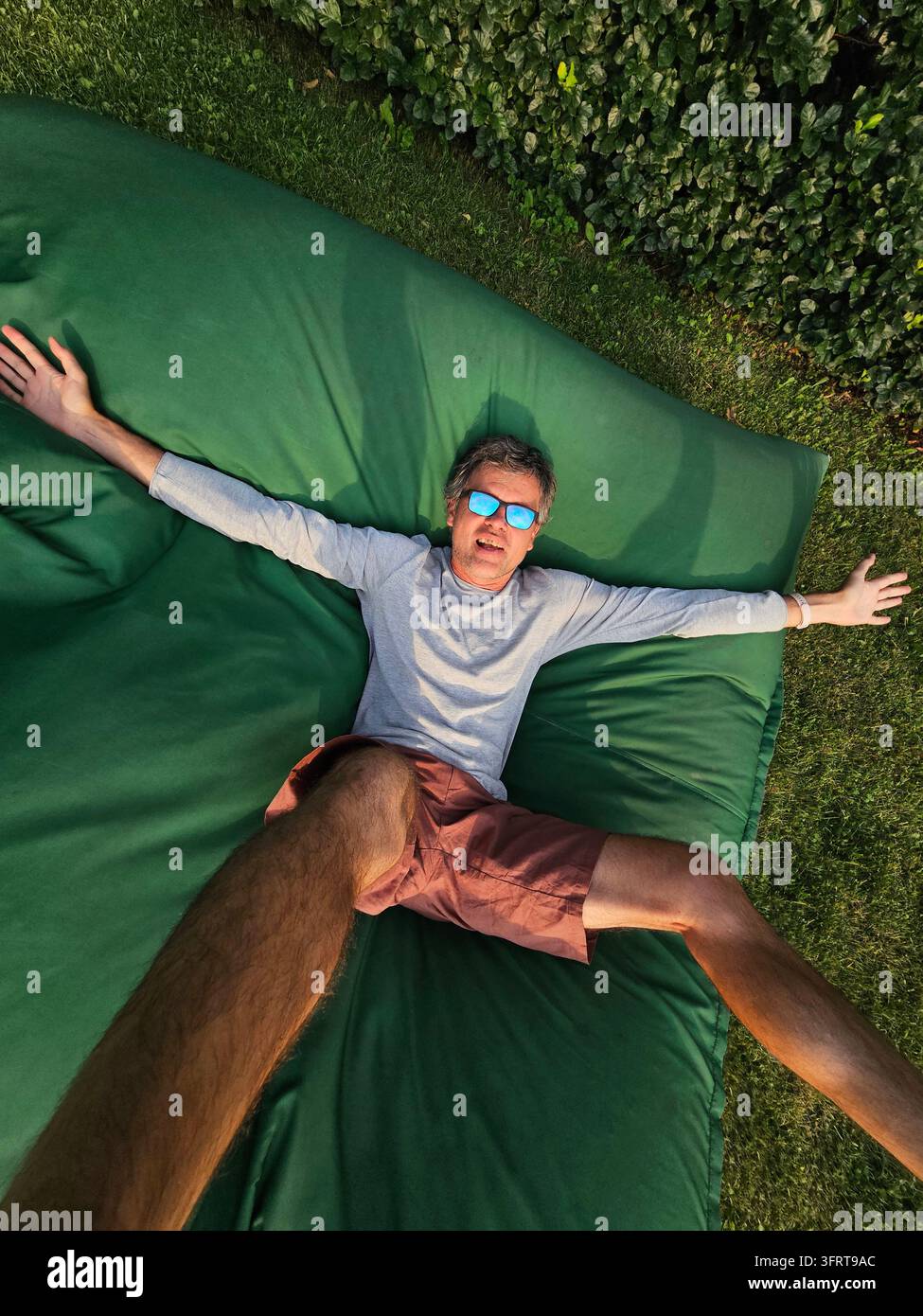 Un homme est allongé sur une chaise verte avec les bras étendus. Il porte des lunettes de soleil et une chemise grise Banque D'Images