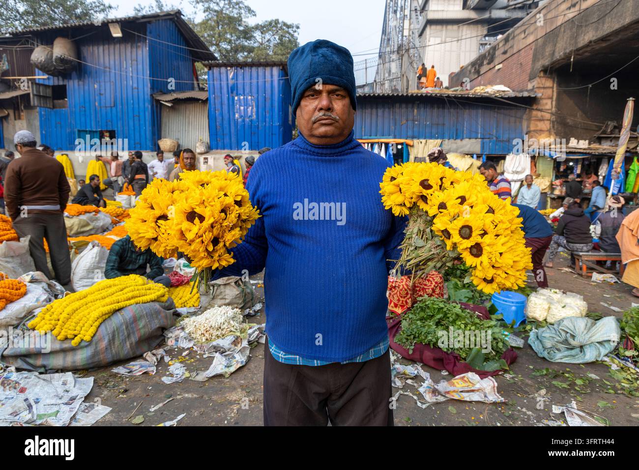 Un homme portant un chapeau de laine bleu se tient les bras écartés tenant des bouquets de tournesols jaunes avec des abris bleus et une foule de commerçants derrière lui au marché aux fleurs Banque D'Images