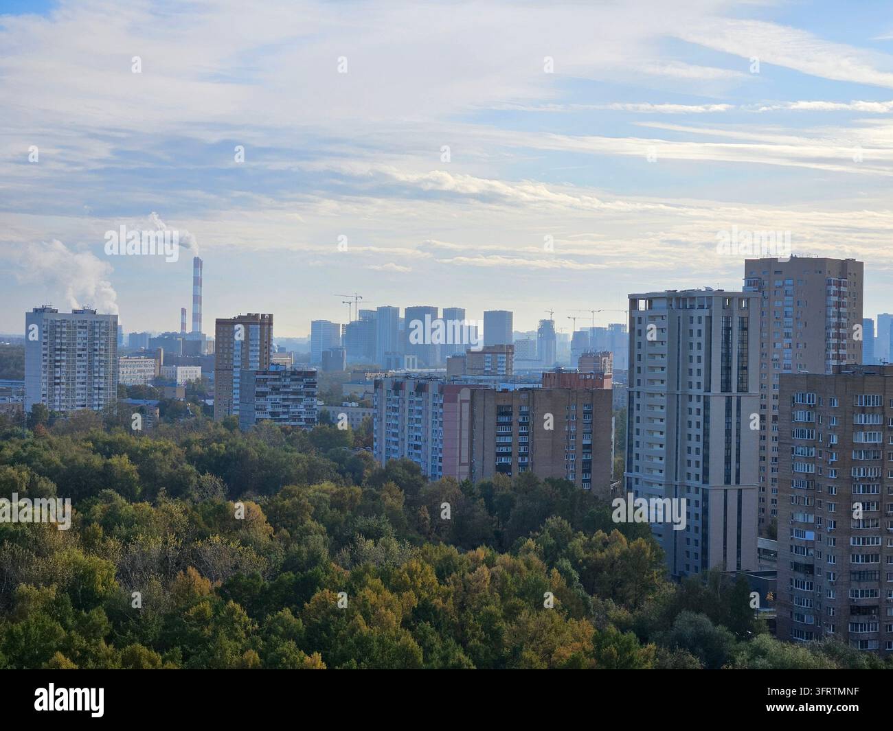 Paysage urbain avec de grands bâtiments résidentiels. Des arbres verts remplissent le premier plan de teintes automnales. Le ciel montre des nuages doux et une cheminée lointaine. Le paysage urbain mêle nature et architecture Banque D'Images