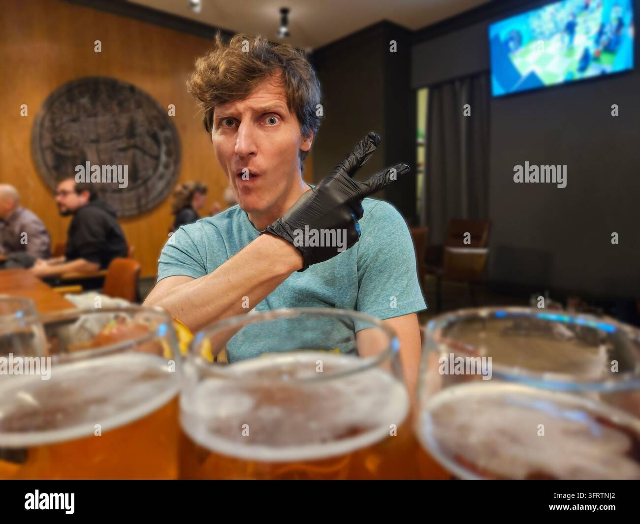 Un homme gesticule avec une main gantée vers des verres de bière. Il est assis à une table dans un cadre de pub. L'arrière-plan montre des clients et un grand emblème rond. Les verres de bière remplissent le premier plan, Banque D'Images