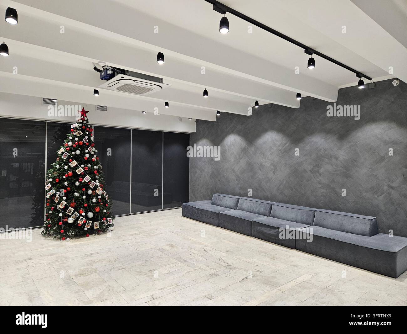 Pièce minimaliste moderne avec arbre de Noël et canapé. Les murs en béton sombre contrastent avec le plafond et le sol blancs. L'éclairage linéaire met en valeur l'espace aux lignes épurées et sobres Banque D'Images
