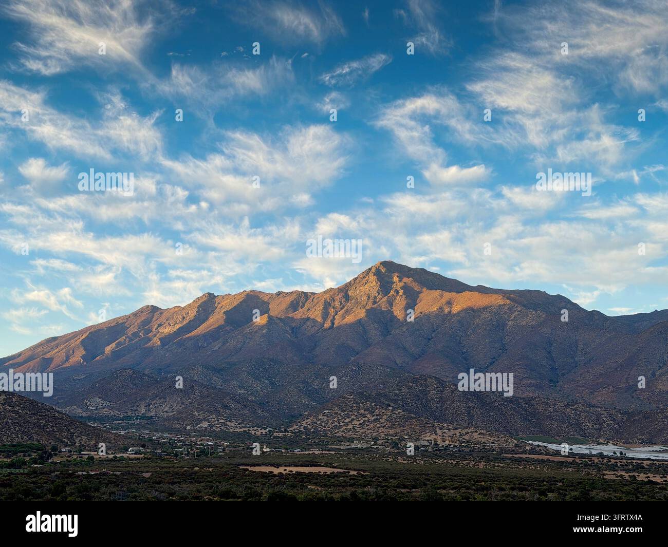 La lumière du soleil illumine le pic escarpé du Cerro La Campana dominant une vallée rurale parsemée d'arbres sous des nuages filamenteux à Olmue Banque D'Images