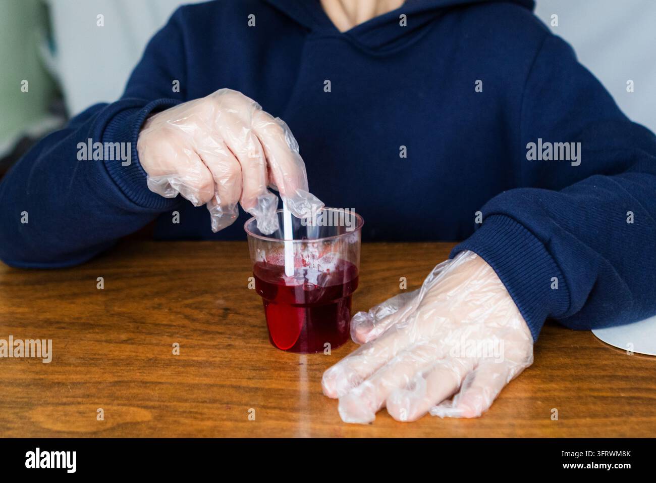 Person portant des gants jetables en train de remuer une boisson rouge dans un gobelet en plastique sur une table en bois Banque D'Images