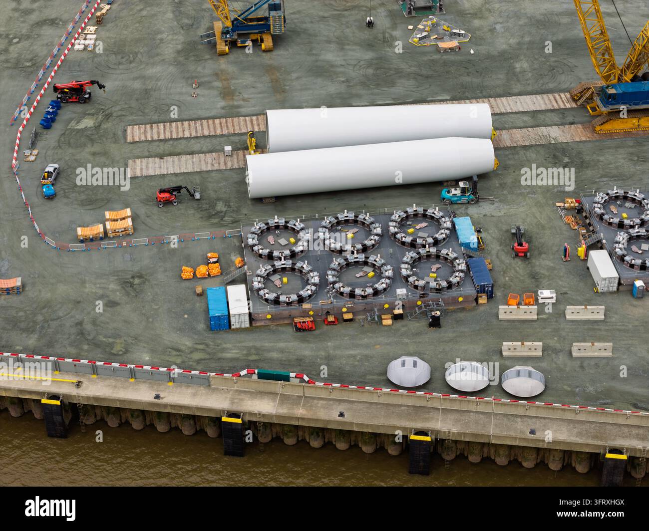 Vue aérienne de grandes sections de tour blanches et de pièces mécaniques de nacelle disposées sur une zone portuaire en béton, prêtes pour le transport vers des parcs éoliens offshore Banque D'Images