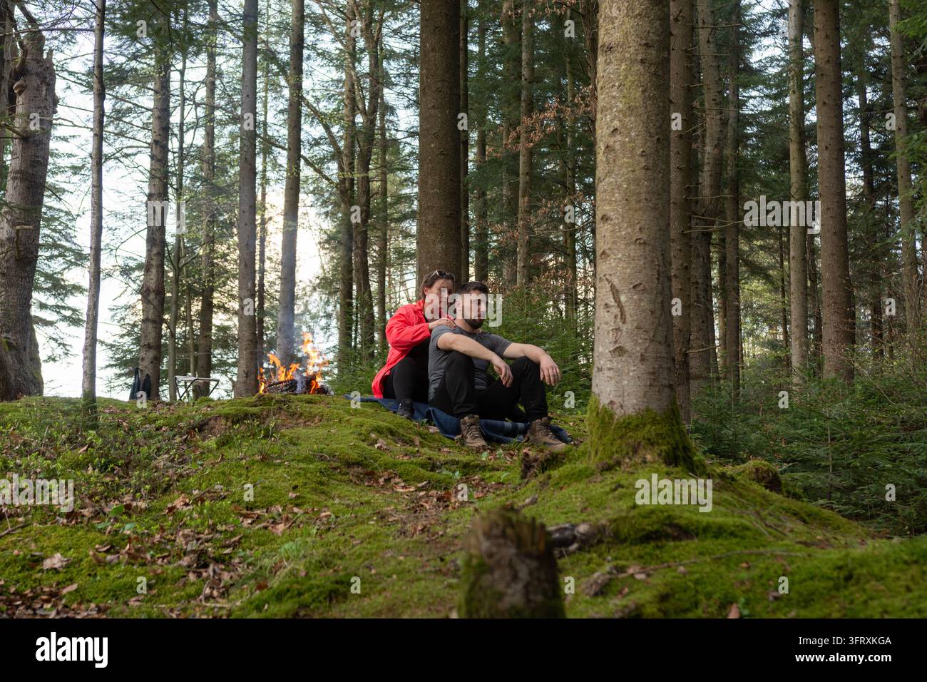 Dans une forêt tranquille, un couple se détend sur de la mousse douce tandis qu'un feu de camp chaud crépite à proximité, entouré de grands arbres et de teintes automnales projetant une lueur dorée Banque D'Images