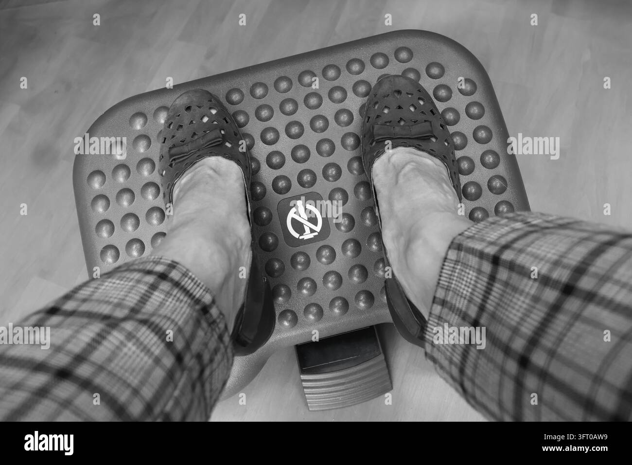 Une photographie monochrome en vue plongeante montre des pieds chaussés de mocassins noirs perforés et d'un pantalon à carreaux reposant sur un repose-pieds surélevé et texturé. La circulaire Banque D'Images