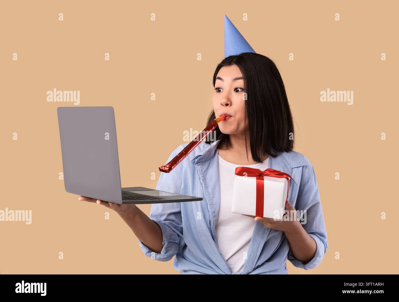 Concept de salutations en ligne. Drôle jeune femme asiatique portant chapeau, tuyau de corne de fête soufflant, tenant l'ordinateur portable et boîte-cadeau enveloppé avec cadeau faire en ligne Banque D'Images