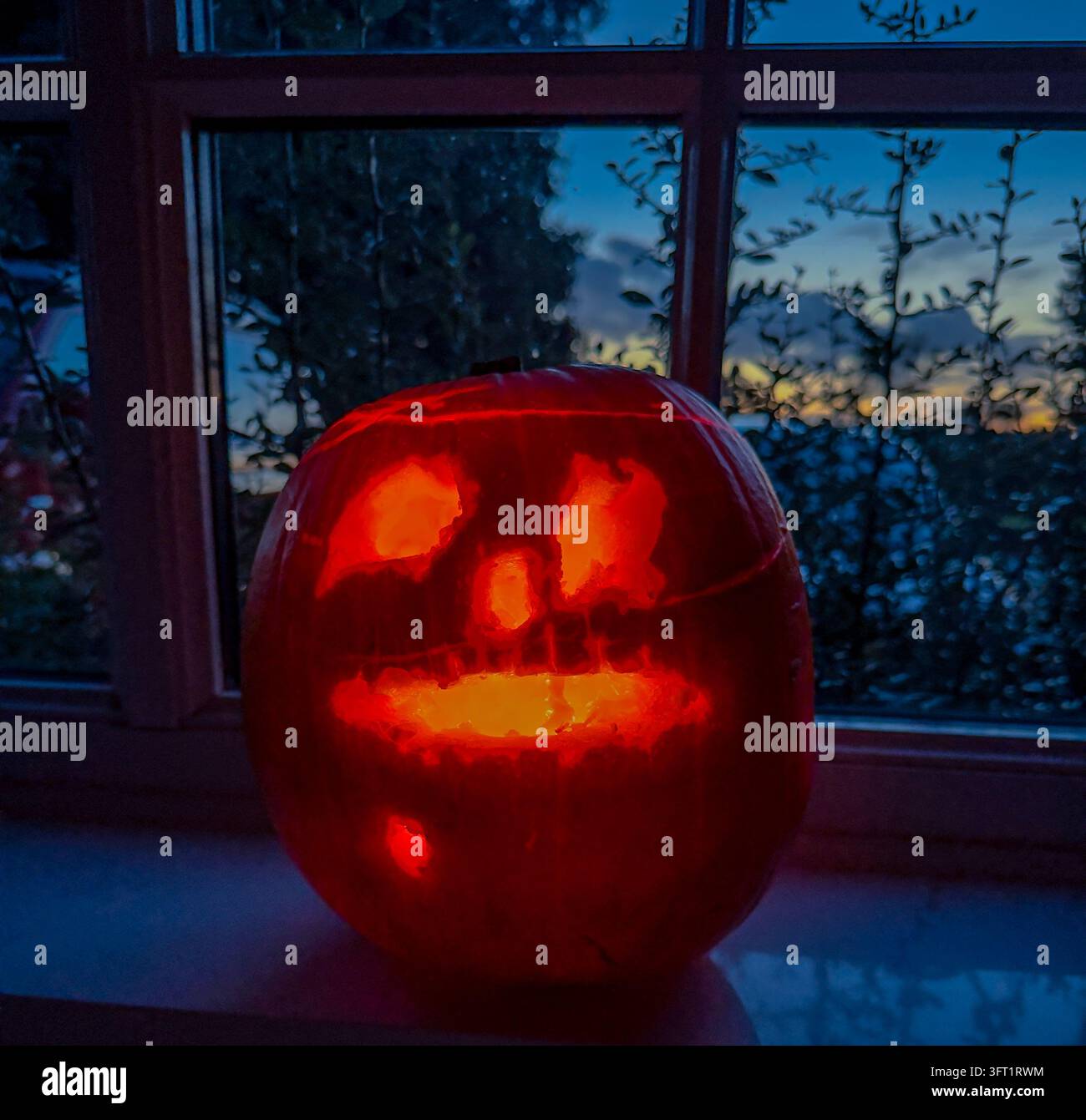 Jack-o’-lanterne d’Halloween sculpté avec un visage surpris lumineux sur un rebord de fenêtre, avec un ciel bleu au crépuscule et des arbres en silhouette en arrière-plan. Banque D'Images