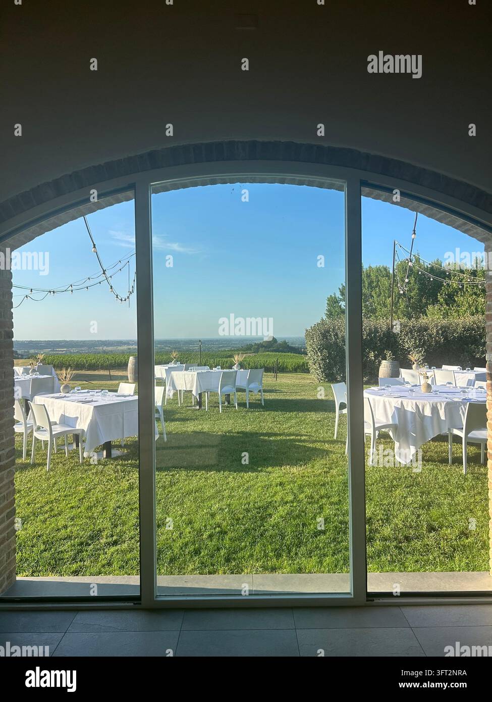 Vue à travers une grande fenêtre en arche de la mise en place d'une réception de mariage en plein air avec des tables de banquet blanches sur une pelouse verte surplombant une campagne pittoresque. - Image de stock capturée avec un smartphone