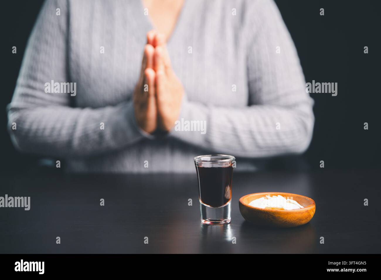 Person en prière derrière des hosties de communion et un petit verre de boisson rouge foncé sur une table noire, illustrant le culte chrétien, la dévotion silencieuse, la foi Banque D'Images