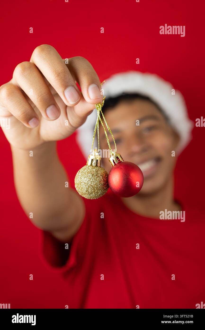 Adolescent à la peau foncée flou tenant deux décorations de Noël, une rouge et une dorée, sur un fond rouge. Image festive joyeuse idéale pour d Banque D'Images
