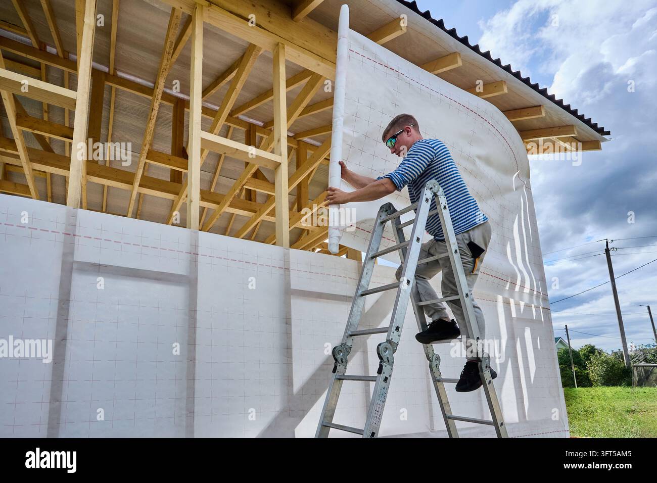 Le revêtement de bâtiment est déroulé par un ouvrier du bâtiment sur le cadre mural en bois pour offrir une protection contre les intempéries et réduire l'infiltration d'air dans une maison à charpente Banque D'Images