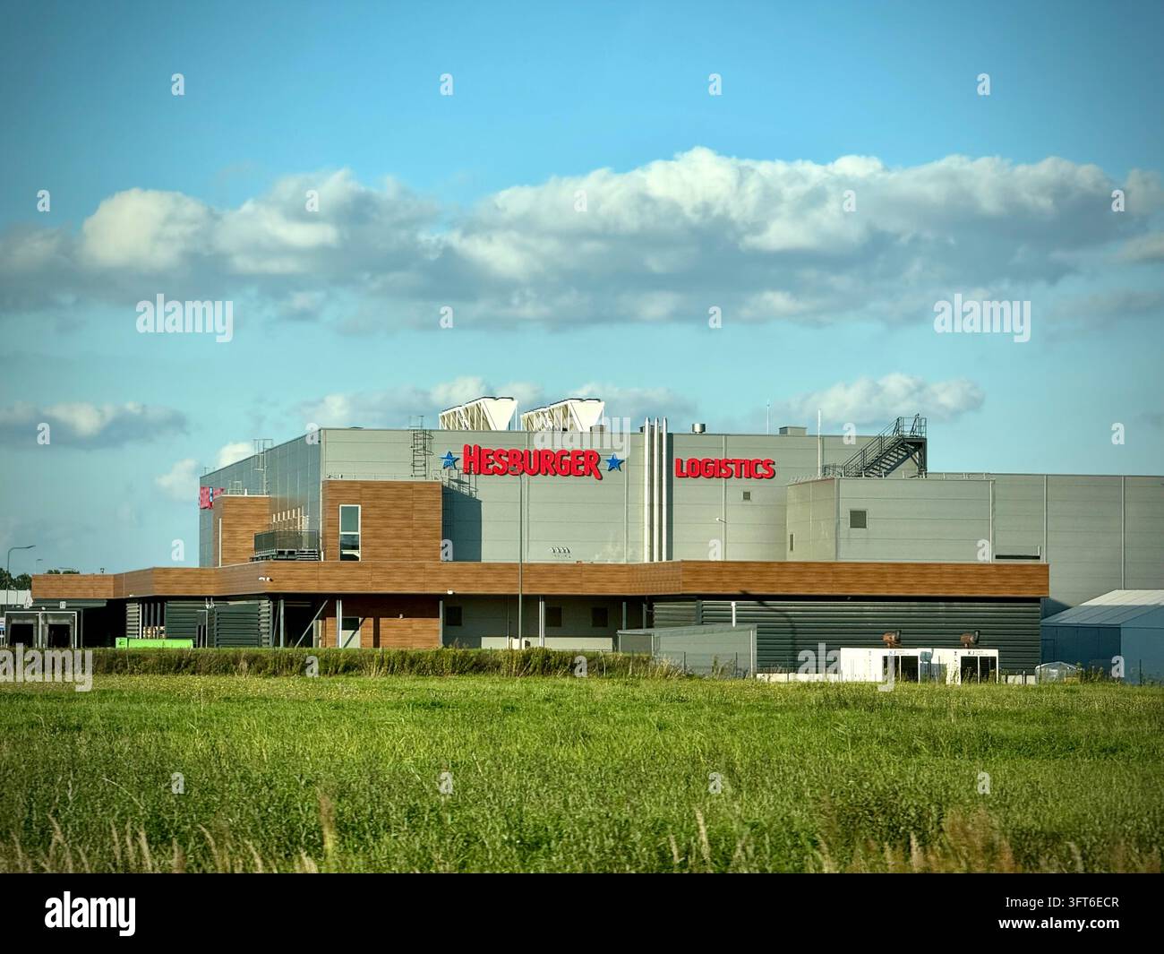 Grand centre de distribution et entrepôt moderne de Hesburger Logistics devant un champ vert et un ciel bleu parsemé de nuages blancs et duveteux. Banque D'Images