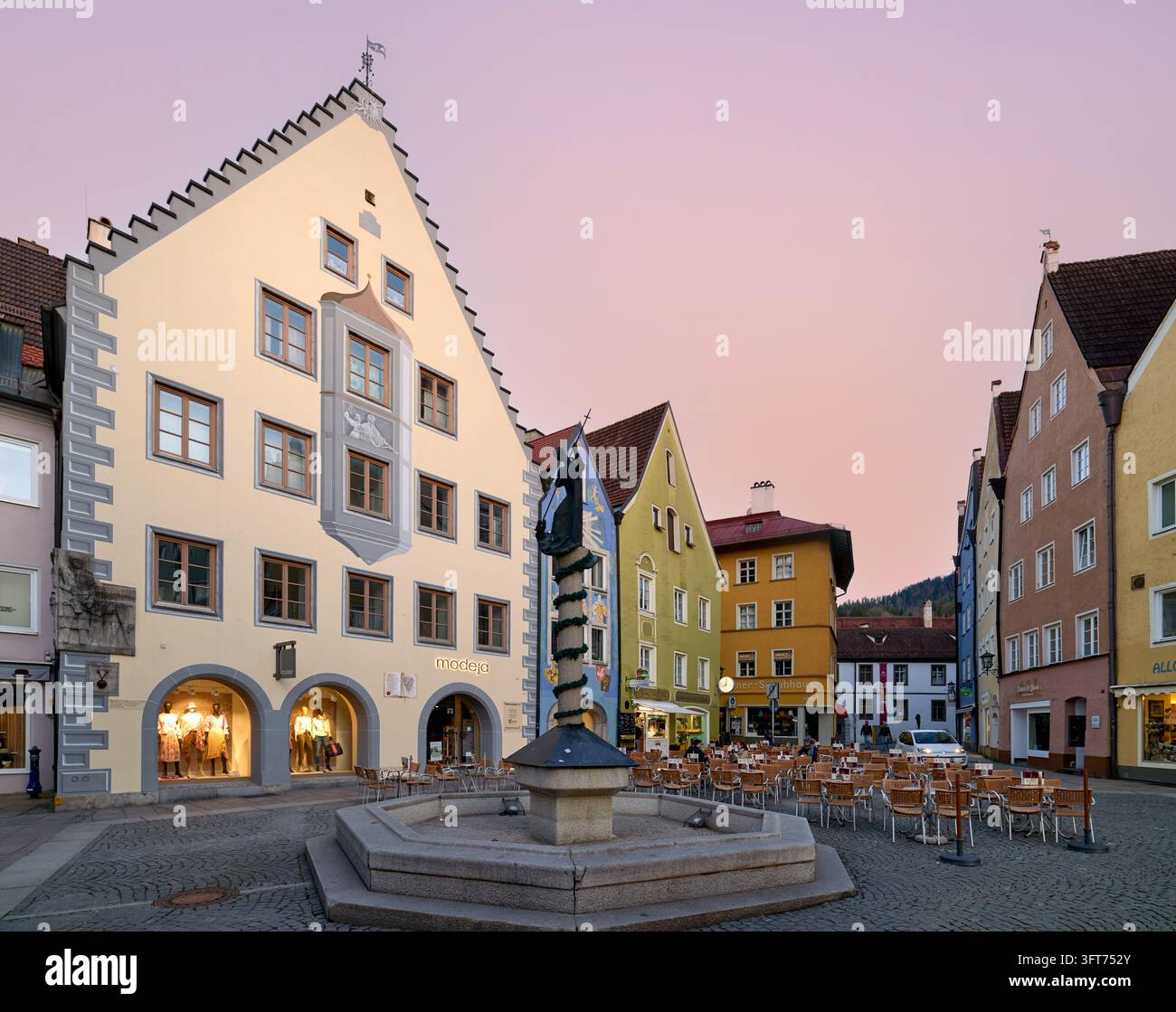 Façade à pignon étagé avec éperon peint, fontaine ornée d'une statue et tables de café vides dans le centre historique de Füssen, en Bavière, sous un ciel rose Banque D'Images