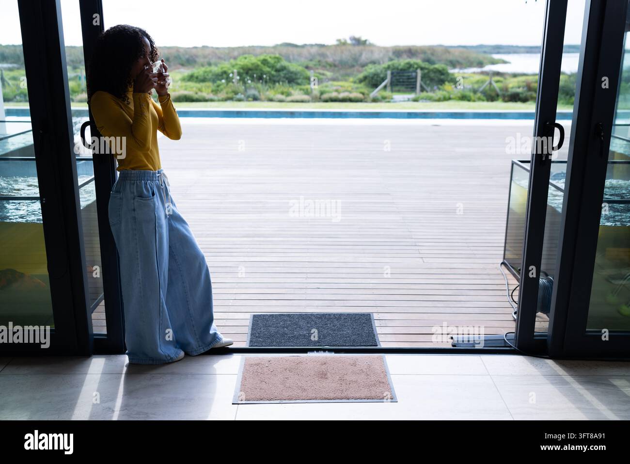 Femme afro-américaine debout près d'une porte coulissante en haut jaune tenant un verre en regardant la piscine Banque D'Images