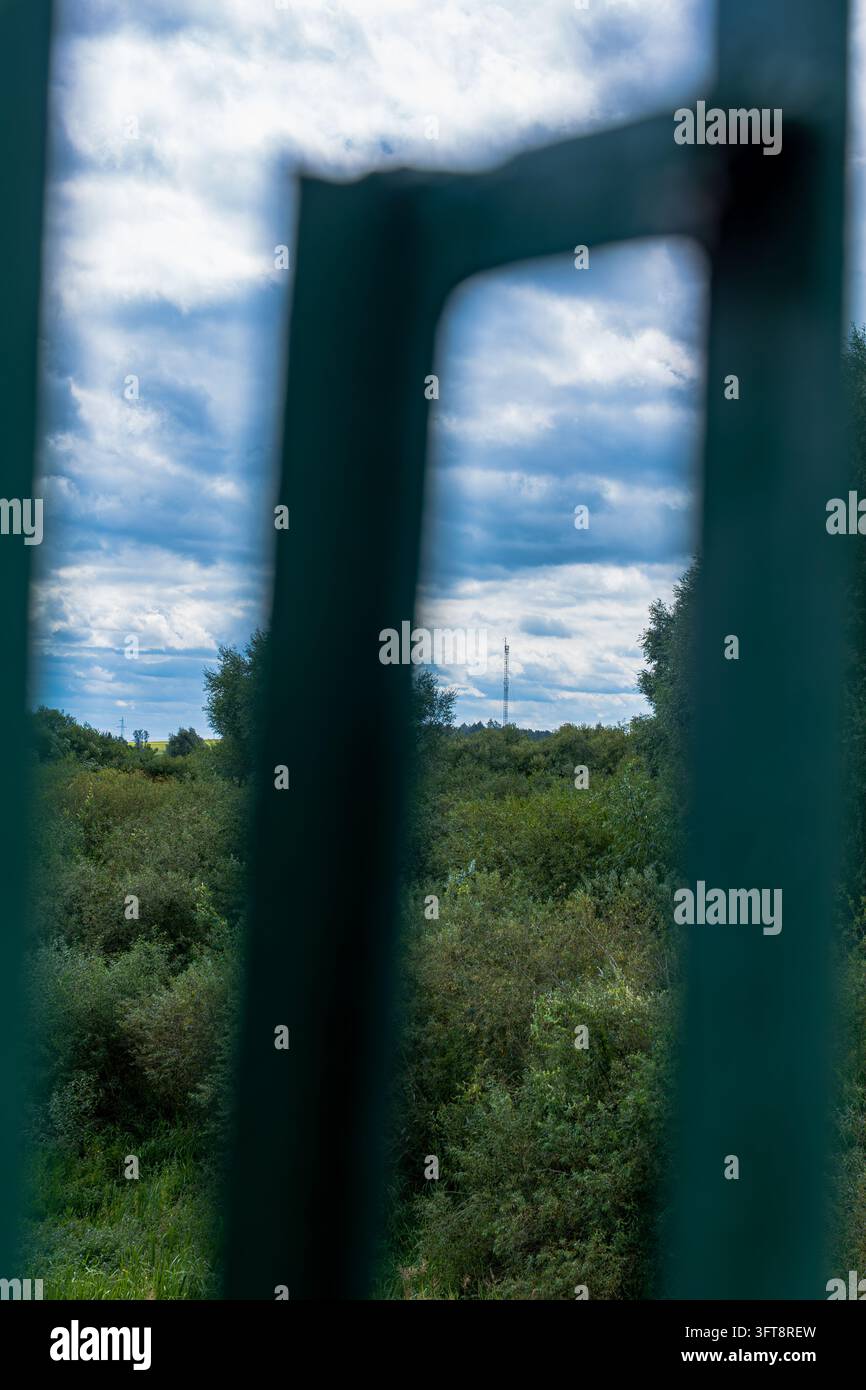 Vue panoramique encadrée à travers une structure métallique industrielle verte montrant un terrain boisé, une tour de communication au loin et un ciel nuageux. Banque D'Images
