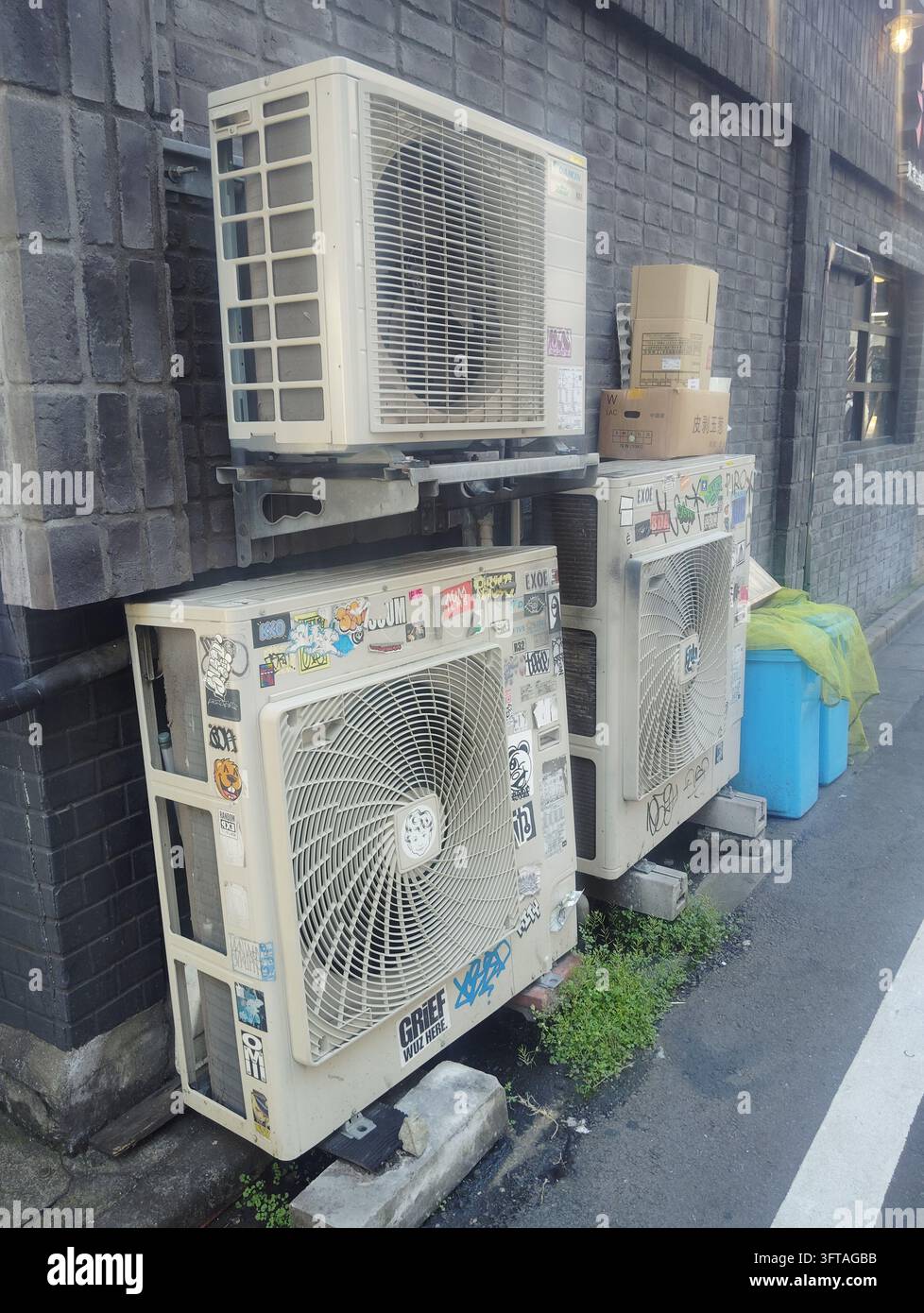 Unités extérieures de climatisation recouvertes d'autocollants de graffitis à côté d'un bâtiment en brique sombre sur une rue de Tokyo, Japon. - Image de stock capturée avec un smartphone