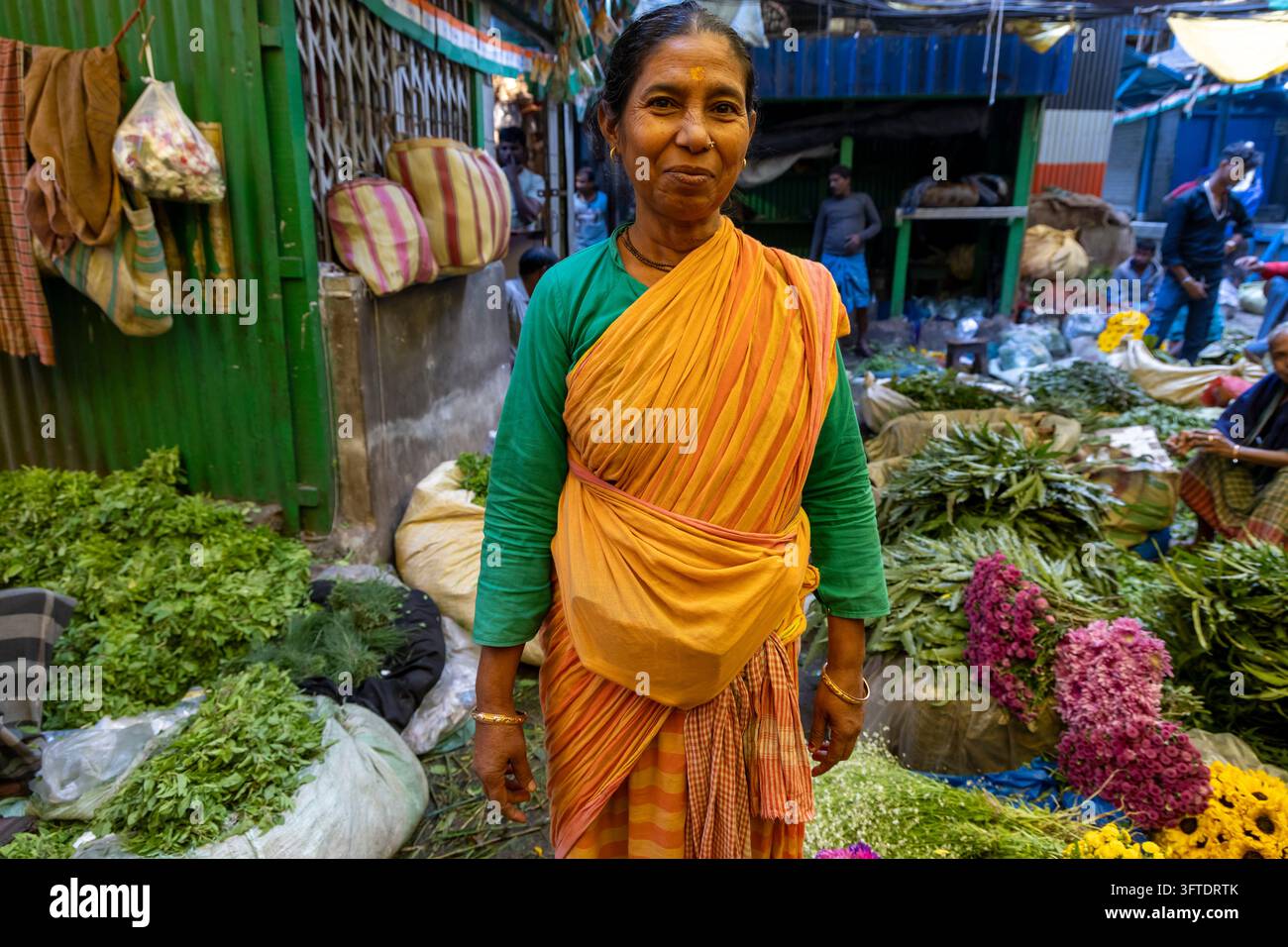 Portrait d'une femme en chemisier vert et tissu orange debout parmi des bouquets feuillus et des sacs au marché aux fleurs, Bengale occidentale, Kolkata, Inde Banque D'Images