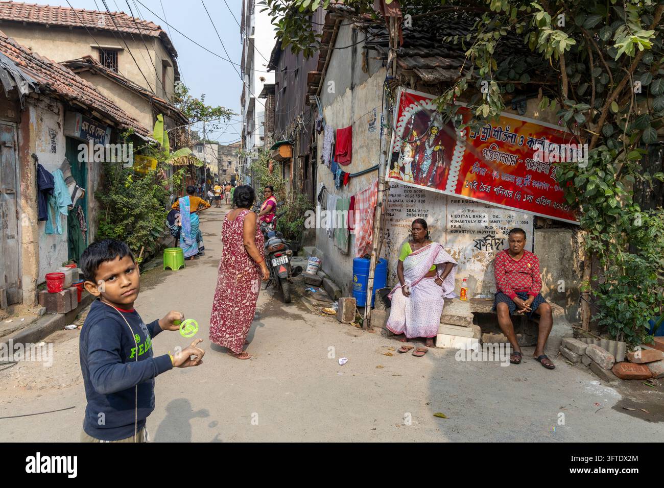 Rue de quartier avec un garçon tenant un jouet à ficelle, une femme en robe rouge marchant et une femme et un homme assis, Bengale occidentale, Kolkata, Inde Banque D'Images