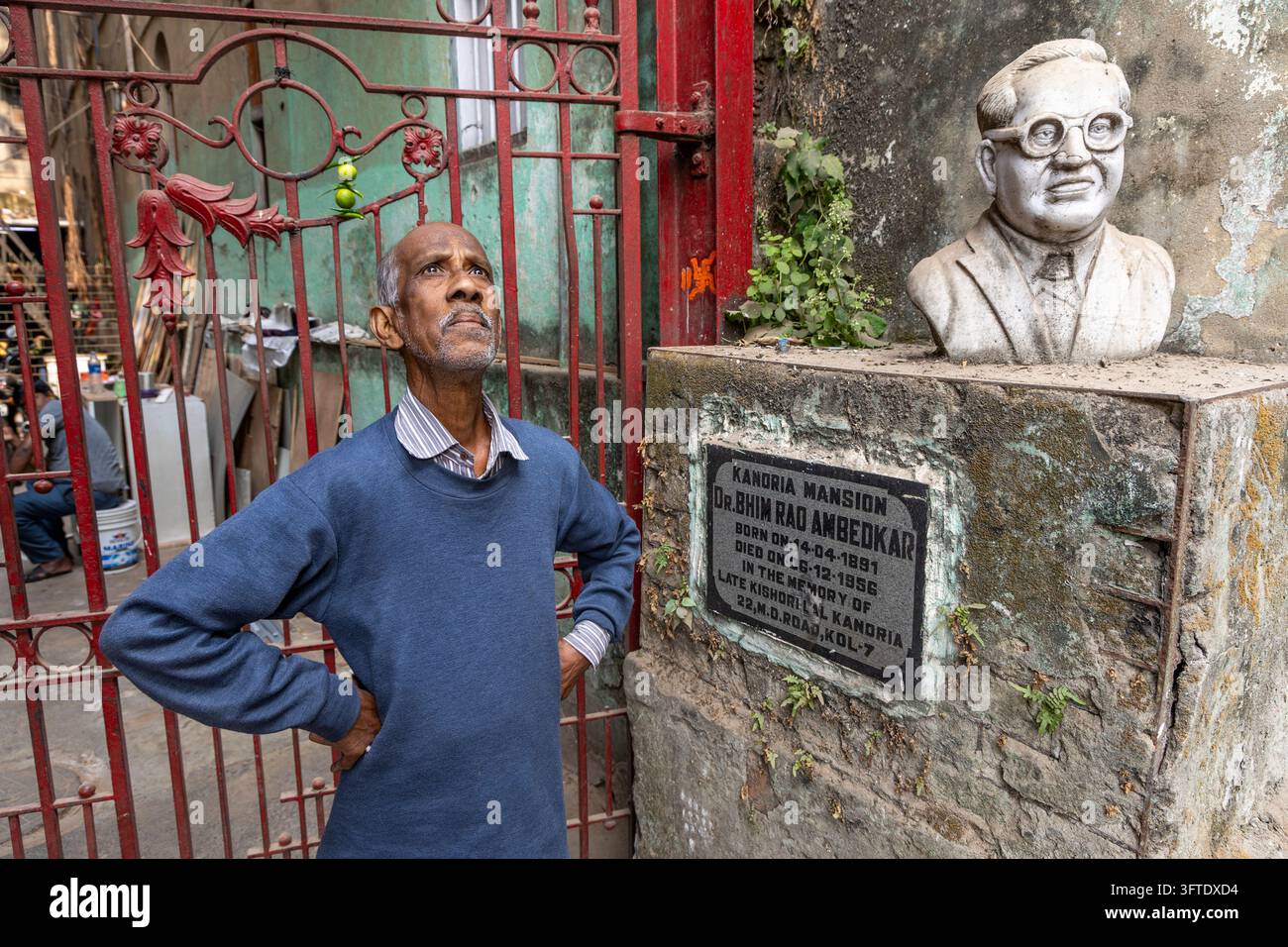 Ancien homme debout à côté d'un monument avec un buste blanc d'un homme à lunettes près d'une porte en fer rouge ornée, Bengale occidentale, Kolkata, Inde Banque D'Images