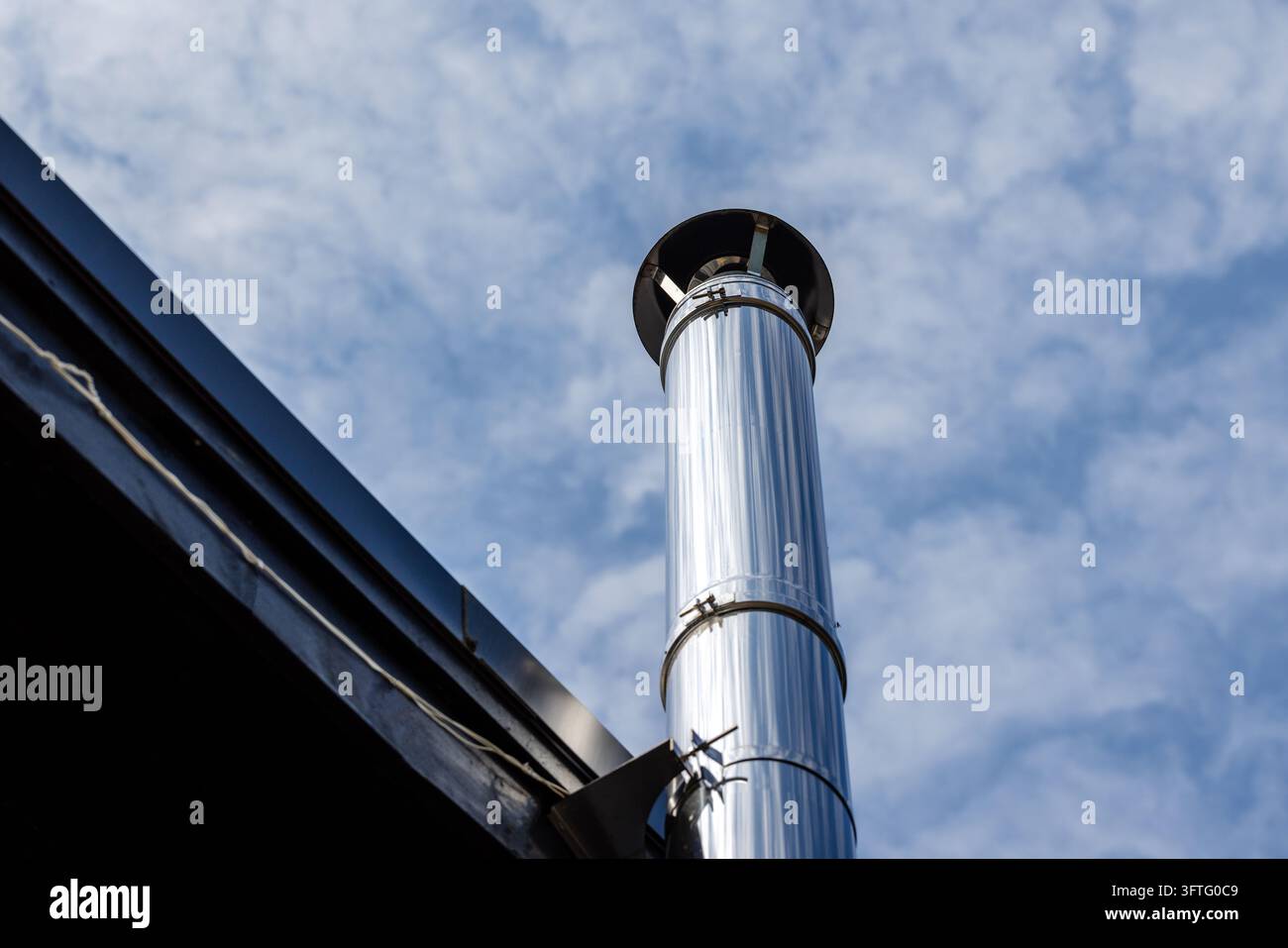 Un conduit de cheminée en acier inoxydable brillant s'élève d'un toit sombre vers un ciel bleu éclatant parsemé de nuages doux, mettant en valeur les détails extérieurs modernes, industri Banque D'Images
