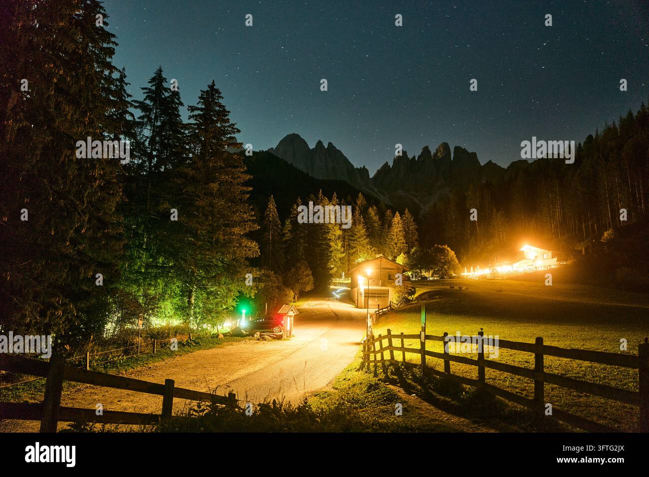 Nuit étoilée sur une route alpine et chalets illuminés sous les pics déchiquetés des Dolomites dans une scène de vallée avec clôture en bois et éclairage de forêt de pins Banque D'Images