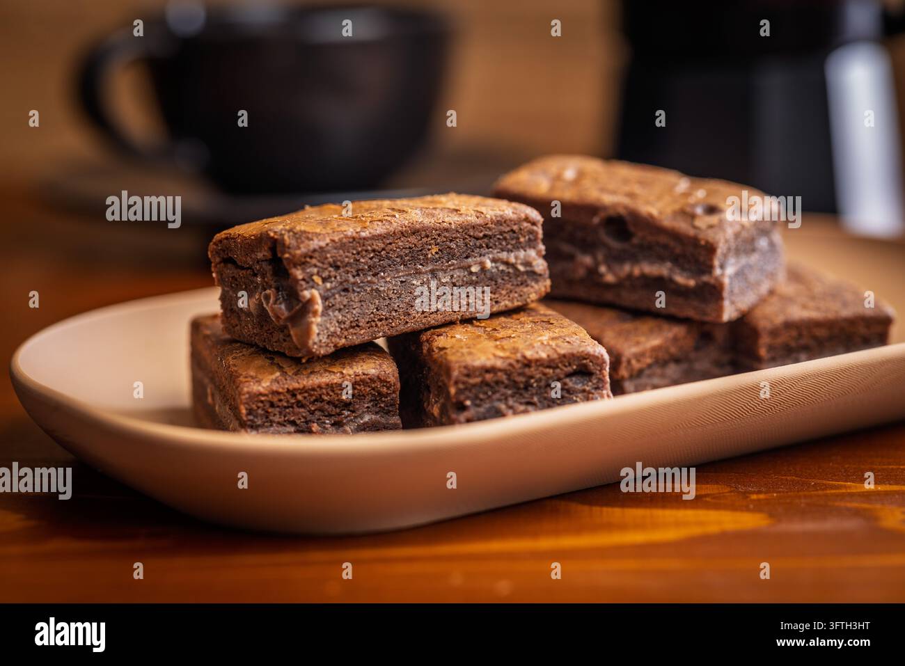 Brownies au chocolat fondants et empilés servis sur une assiette beige avec une tasse de café floue en arrière-plan. Photographie culinaire chaleureuse et confortable mettant en valeur la texture Banque D'Images