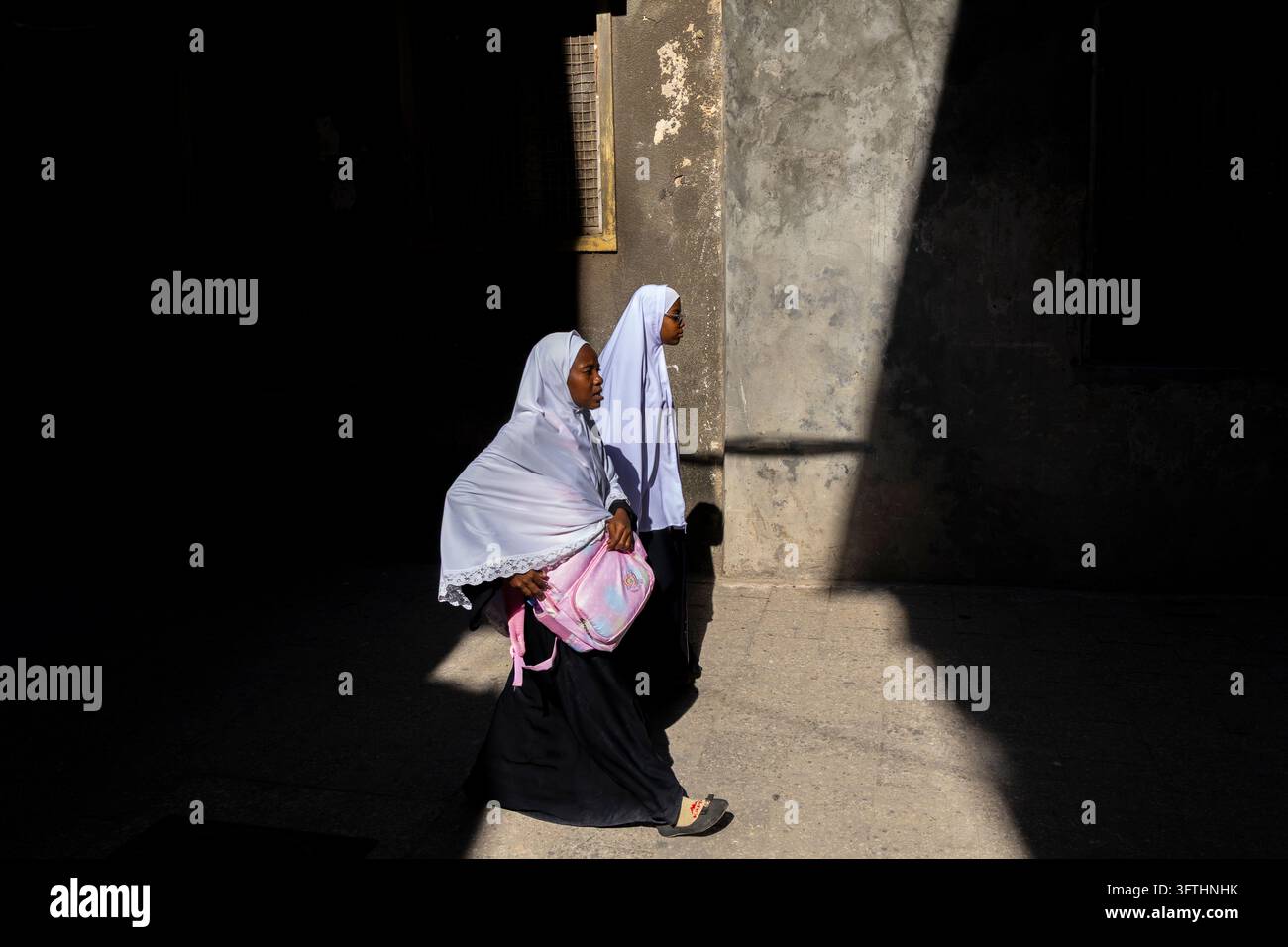 Deux écolières en hijabs blancs et robes noires, l'une avec un sac à dos rose, marchant à travers la lumière du soleil et les ombres profondes, Stone Town, Tanzanie Banque D'Images