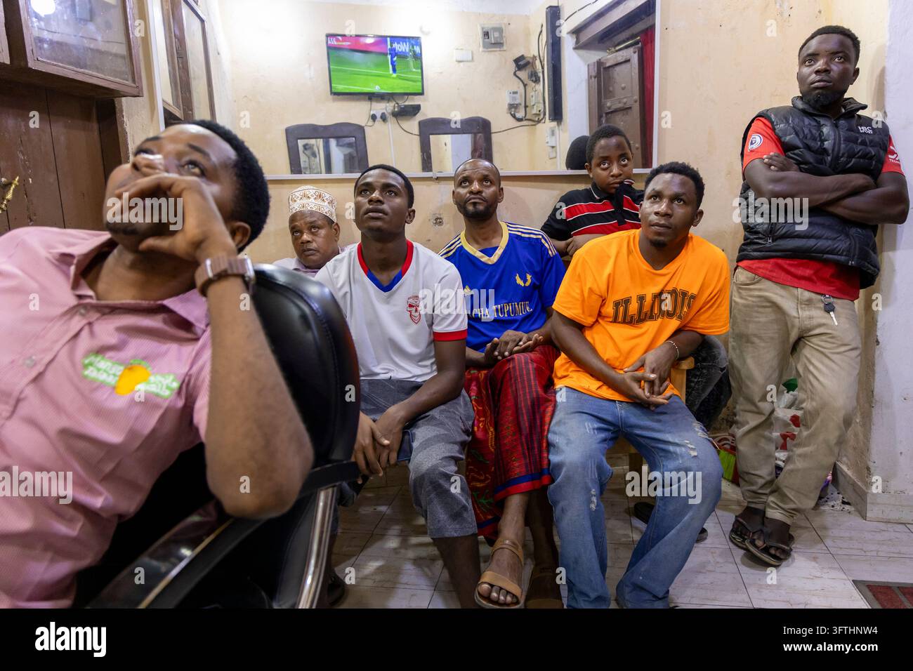 Hommes et un garçon regardant le football à la télévision dans un petit salon de coiffure avec un écran plat murale et un miroir, Stone Town, Tanzanie Banque D'Images