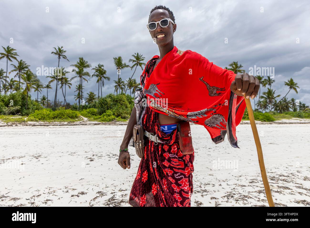 Homme souriant de la tribu Maasai portant des lunettes de soleil et un shuka rouge avec motif girafe et zèbre, appuyé sur un bâton sur une plage de sable blanc à Matemwe, Tanzanie Banque D'Images