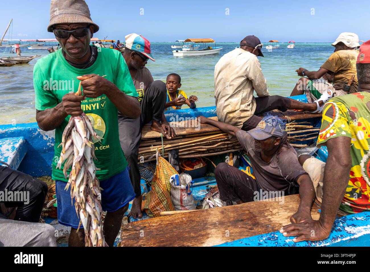 Pêcheurs dans un bateau en bois bleu triant leur prise tandis qu'un homme en chemise verte tient de petits poissons avec des bateaux de touristes en arrière-plan, Matemwe, Tanzanie Banque D'Images