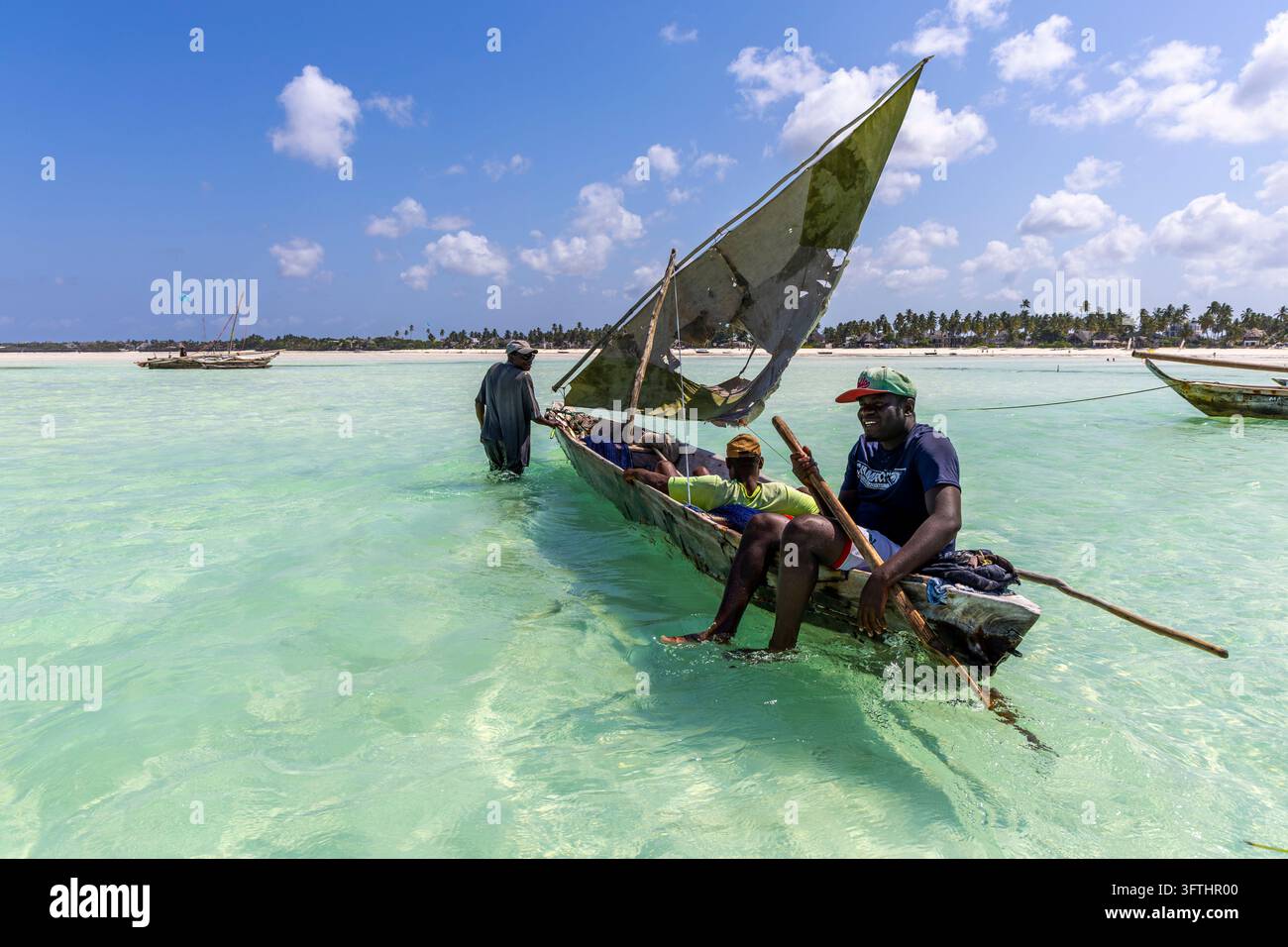 Pêcheurs dans une pirogue à balancier en bois avec une voile déchirée dans une eau turquoise peu profonde avec un homme assis tenant une perche, Jambiani, Tanzanie Banque D'Images