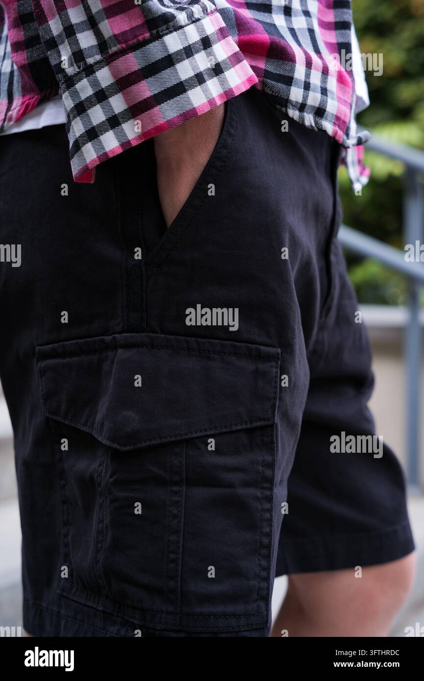 Un jeune homme méconnaissable, vêtu d'un t-shirt blanc, d'une chemise à carreaux rose et d'un short cargo noir, se tient la main dans la poche en plein air Banque D'Images