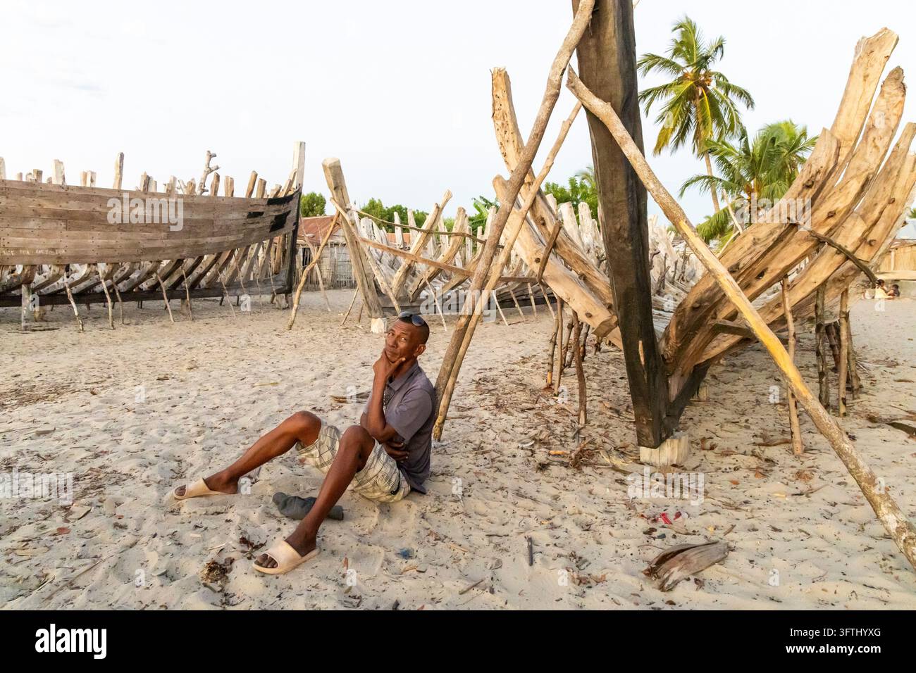 Homme Vezo assis sur le sable, reposant contre des carcasses de bateaux en bois dans une chantier naval animé, Menabe, Belo sur Mer, Madagascar Banque D'Images