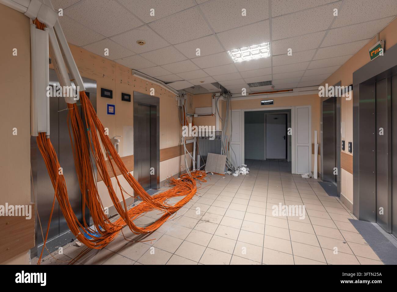 Des faisceaux de câbles orange pendent d'un conduit de câbles ouvert dans un hall d'ascenseur avec des portes métalliques et des débris sur le sol Banque D'Images