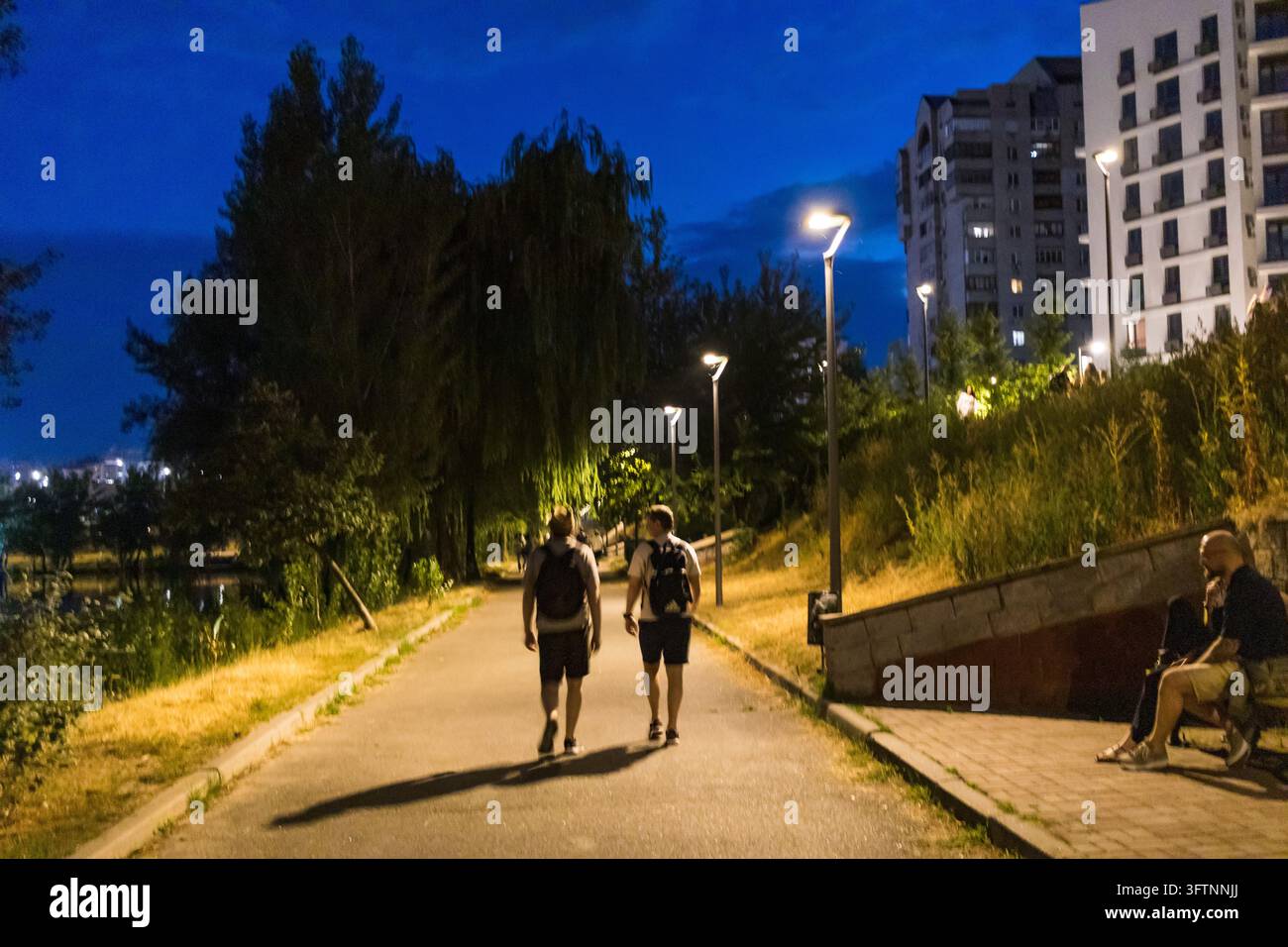 Des amis se promènent le long d'une promenade lacustre sous les lampadaires à l'heure bleue tandis que d'autres se reposent sur des bancs, soirée paisible à Kyiv Banque D'Images